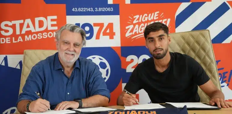IMG_8440 | AllezPaillade.com Two men sign documents at a table during a signing event, with a bright orange–blue graphic wall behind them.