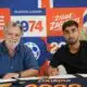 Two men sign documents at a table during a signing event, with a bright orange&ndash;blue graphic wall behind them.