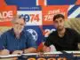 Two men sign documents at a table during a signing event, with a bright orange&ndash;blue graphic wall behind them.