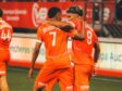 Two orange-clad soccer players embrace on the field, jersey numbers 7 and 9 visible, as a teammate stands in the background.