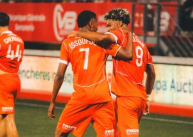 Issoufou Megnan | AllezPaillade.com Two orange-clad soccer players embrace on the field, jersey numbers 7 and 9 visible, as a teammate stands in the background.