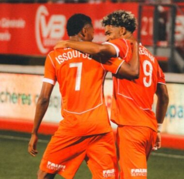 Two orange-clad soccer players embrace on the field, jersey numbers 7 and 9 visible, as a teammate stands in the background.