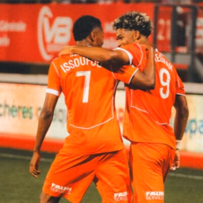 Two orange-clad soccer players embrace on the field, jersey numbers 7 and 9 visible, as a teammate stands in the background.