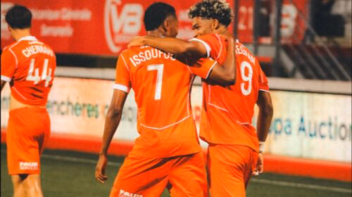 Two orange-clad soccer players embrace on the field, jersey numbers 7 and 9 visible, as a teammate stands in the background.