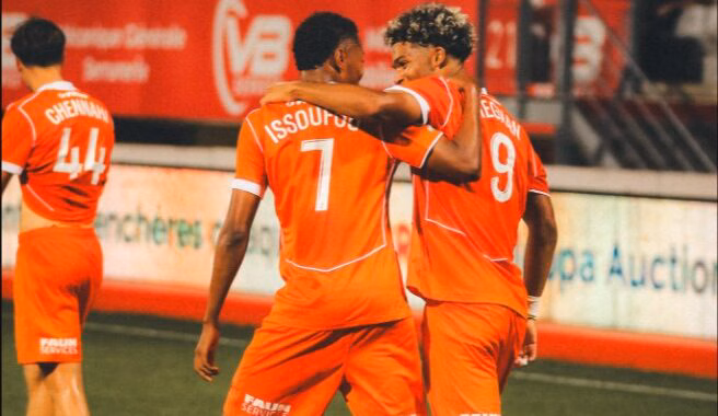 Two orange-clad soccer players embrace on the field, jersey numbers 7 and 9 visible, as a teammate stands in the background.