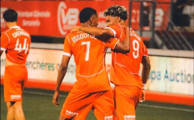 Two orange-clad soccer players embrace on the field, jersey numbers 7 and 9 visible, as a teammate stands in the background.