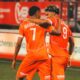 Two orange-clad soccer players embrace on the field, jersey numbers 7 and 9 visible, as a teammate stands in the background.