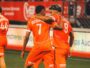 Two orange-clad soccer players embrace on the field, jersey numbers 7 and 9 visible, as a teammate stands in the background.