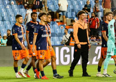 Group of soccer players in navy blue and orange kits walking on a grass pitch, stadium seating in the background.