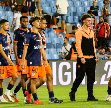 Group of soccer players in navy blue and orange kits walking on a grass pitch, stadium seating in the background.