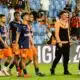 Group of soccer players in navy blue and orange kits walking on a grass pitch, stadium seating in the background.