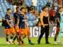 Group of soccer players in navy blue and orange kits walking on a grass pitch, stadium seating in the background.