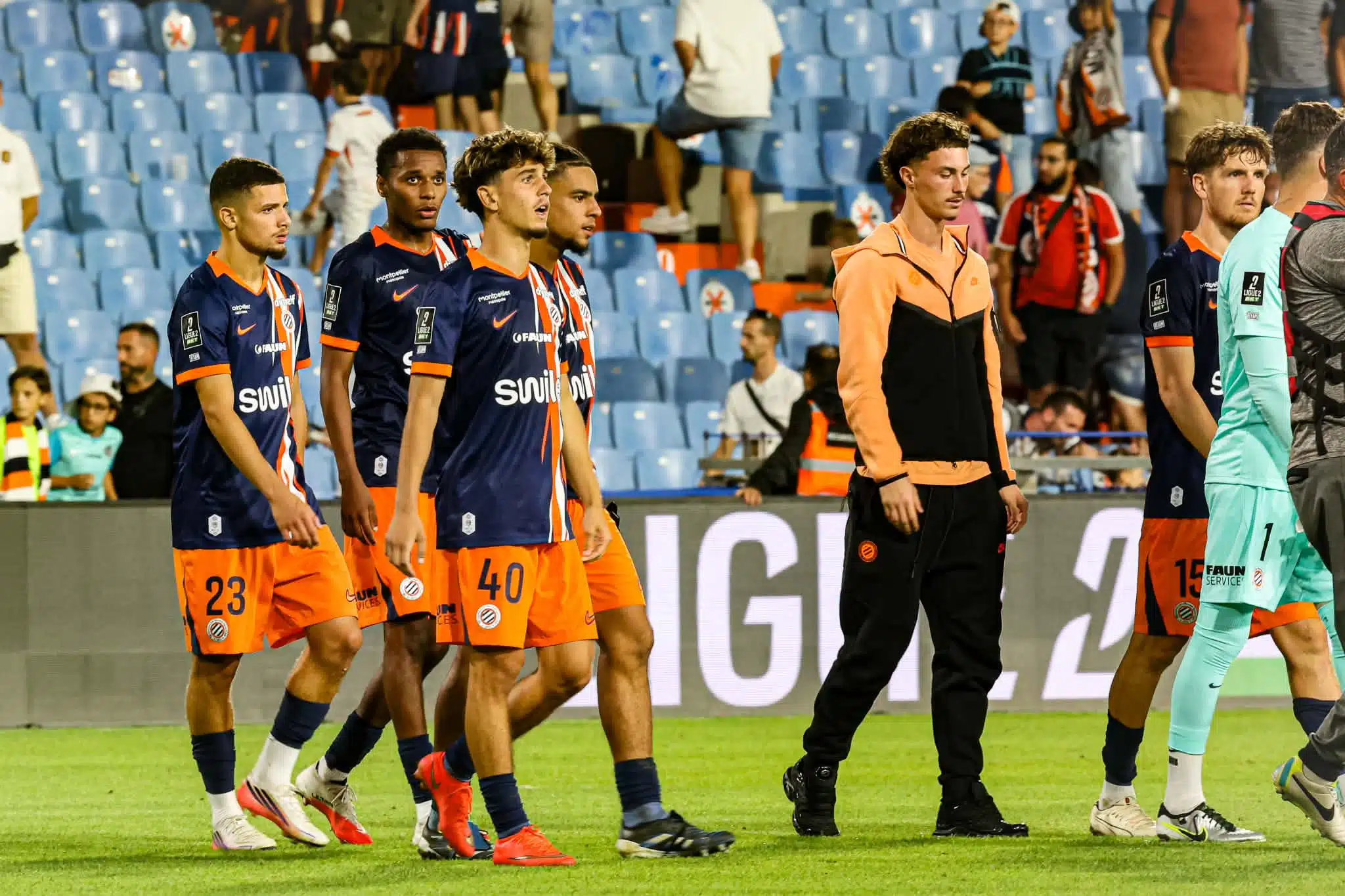 Group of soccer players in navy blue and orange kits walking on a grass pitch, stadium seating in the background.