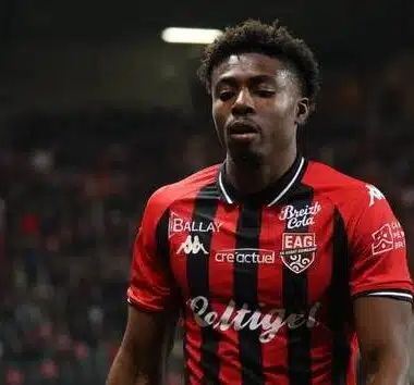 Male soccer player in a red-and-black striped jersey on a green pitch with a blurred crowd in the background.