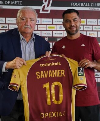 Two men hold up a maroon and yellow soccer jersey reading 'SAVANIER' and number 10, in front of a Metz FC sponsor wall.
