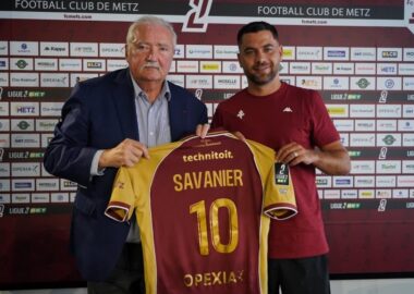 Two men hold up a maroon and yellow soccer jersey reading 'SAVANIER' and number 10, in front of a Metz FC sponsor wall.
