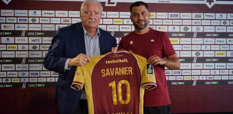 Savanier | AllezPaillade.com Two men hold up a maroon and yellow soccer jersey reading 'SAVANIER' and number 10, in front of a Metz FC sponsor wall.
