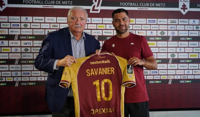 Two men hold up a maroon and yellow soccer jersey reading 'SAVANIER' and number 10, in front of a Metz FC sponsor wall.
