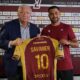 Two men hold up a maroon and yellow soccer jersey reading 'SAVANIER' and number 10, in front of a Metz FC sponsor wall.