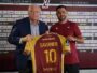 Two men hold up a maroon and yellow soccer jersey reading 'SAVANIER' and number 10, in front of a Metz FC sponsor wall.