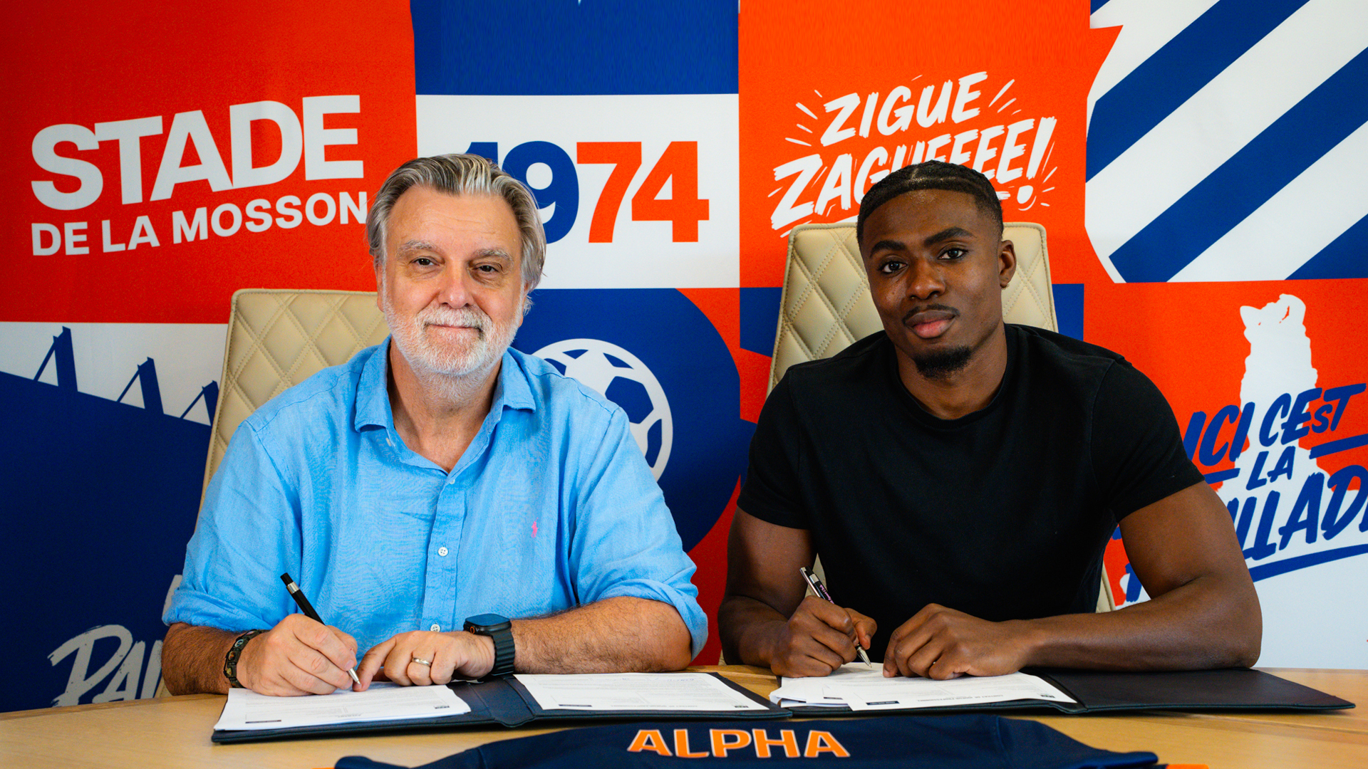 Two men sign documents at a desk in a bright, sports-themed backdrop behind them.
