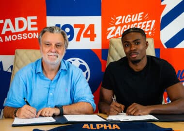 Two men sit at a table signing documents; bright red, blue, and orange mural behind them with 'ALPHA' branding.