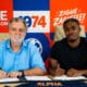 Two men sit at a table signing documents; bright red, blue, and orange mural behind them with 'ALPHA' branding.
