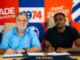 Two men sit at a table signing documents; bright red, blue, and orange mural behind them with 'ALPHA' branding.