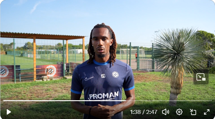 Athlete wearing a navy blue football jersey with PROMAN on the chest, standing on a grassy practice field with a fence and palm plant in the background.