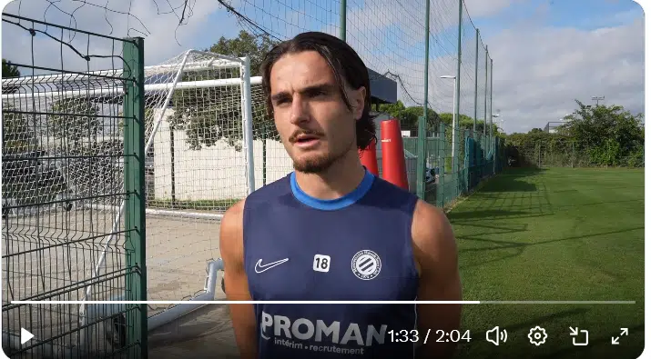 image | AllezPaillade.com Male football player in a blue training kit being interviewed on a sunny training ground beside a wire fence.