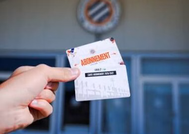 images 26 | AllezPaillade.com A hand holding a subscription card labeled ABONNEMENT in front of a building entrance with a circular emblem above.