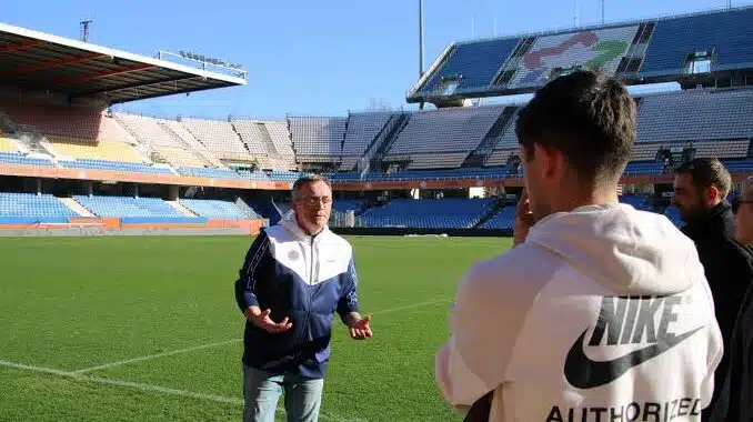 images 8 | AllezPaillade.com Coach on a sunny soccer pitch explaining something to a small group, with empty stadium stands in the background and a man wearing a Nike hoodie nearby.