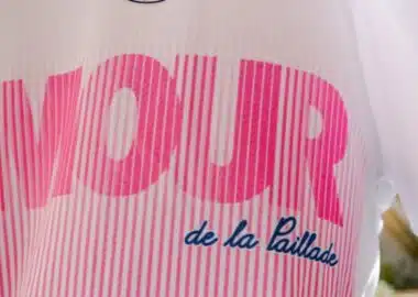 Close-up of a pink striped logo with large bold letters on white fabric; blue cursive text reads 'de la Paillade'.