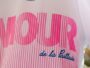 Close-up of a pink striped logo with large bold letters on white fabric; blue cursive text reads 'de la Paillade'.