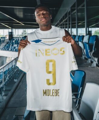 Soccer player stands in a stadium concourse holding up a white jersey with gold lettering, INEOS, and number 9.