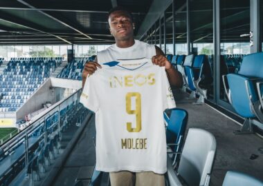 Soccer player stands in a stadium concourse holding up a white jersey with gold lettering, INEOS, and number 9.