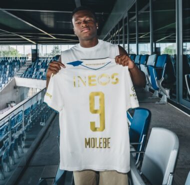 Soccer player stands in a stadium concourse holding up a white jersey with gold lettering, INEOS, and number 9.