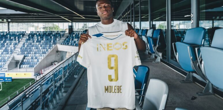 molebe lausanne tribunes | AllezPaillade.com Soccer player stands in a stadium concourse holding up a white jersey with gold lettering, INEOS, and number 9.