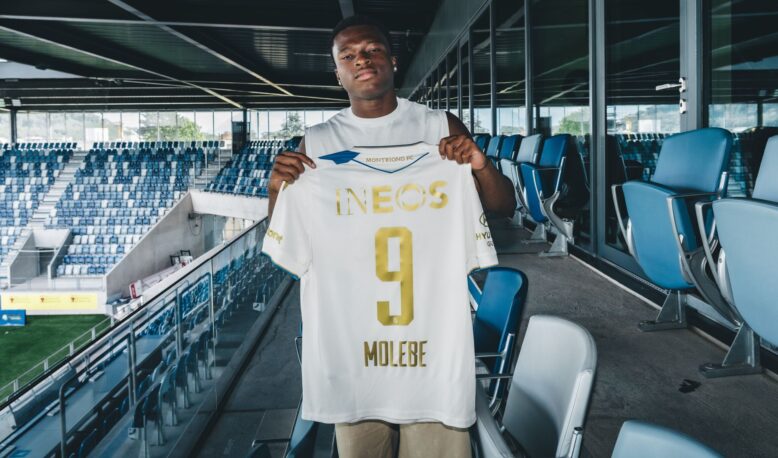 Soccer player stands in a stadium concourse holding up a white jersey with gold lettering, INEOS, and number 9.