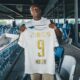 Soccer player stands in a stadium concourse holding up a white jersey with gold lettering, INEOS, and number 9.