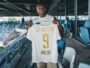 Soccer player stands in a stadium concourse holding up a white jersey with gold lettering, INEOS, and number 9.