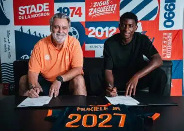 Two men sit at a table signing documents, with a colorful poster backdrop that reads 1974 and 2012 and features bold red, blue, and orange panels behind them. The man on the left wears an orange polo; the man on the right wears a black shirt. A navy and orange jersey on the table in front shows the name 'MUKIELE JR' and '2021'.