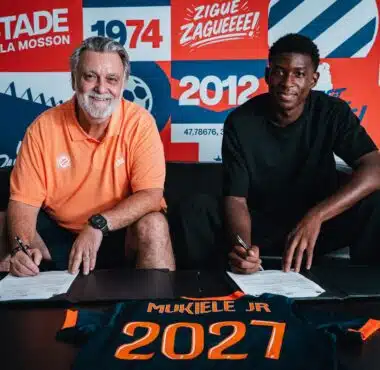 Two men sit at a table signing documents, with a colorful poster backdrop that reads 1974 and 2012 and features bold red, blue, and orange panels behind them. The man on the left wears an orange polo; the man on the right wears a black shirt. A navy and orange jersey on the table in front shows the name 'MUKIELE JR' and '2021'.