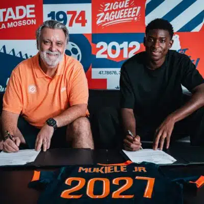 Two men sit at a table signing documents, with a colorful poster backdrop that reads 1974 and 2012 and features bold red, blue, and orange panels behind them. The man on the left wears an orange polo; the man on the right wears a black shirt. A navy and orange jersey on the table in front shows the name 'MUKIELE JR' and '2021'.