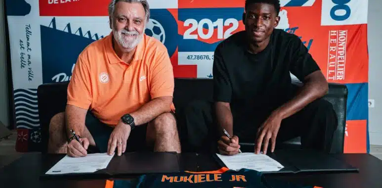mukiele | AllezPaillade.com Two men sit at a table signing documents, with a colorful poster backdrop that reads 1974 and 2012 and features bold red, blue, and orange panels behind them. The man on the left wears an orange polo; the man on the right wears a black shirt. A navy and orange jersey on the table in front shows the name 'MUKIELE JR' and '2021'.