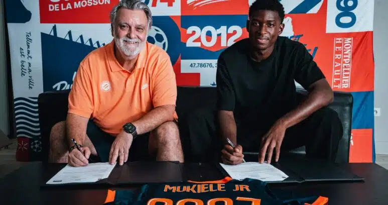 Two men sit at a table signing documents, with a colorful poster backdrop that reads 1974 and 2012 and features bold red, blue, and orange panels behind them. The man on the left wears an orange polo; the man on the right wears a black shirt. A navy and orange jersey on the table in front shows the name 'MUKIELE JR' and '2021'.