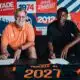 Two men sit at a table signing documents, with a colorful poster backdrop that reads 1974 and 2012 and features bold red, blue, and orange panels behind them. The man on the left wears an orange polo; the man on the right wears a black shirt. A navy and orange jersey on the table in front shows the name 'MUKIELE JR' and '2021'.