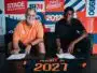 Two men sit at a table signing documents, with a colorful poster backdrop that reads 1974 and 2012 and features bold red, blue, and orange panels behind them. The man on the left wears an orange polo; the man on the right wears a black shirt. A navy and orange jersey on the table in front shows the name 'MUKIELE JR' and '2021'.