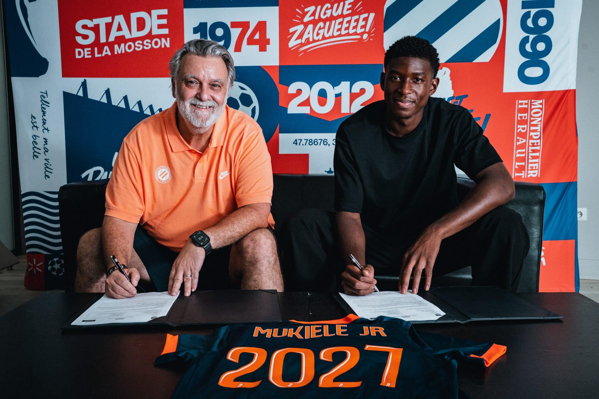 Two men sit at a table signing documents, with a colorful poster backdrop that reads 1974 and 2012 and features bold red, blue, and orange panels behind them. The man on the left wears an orange polo; the man on the right wears a black shirt. A navy and orange jersey on the table in front shows the name'MUKIELE JR' and '2021'.