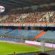 Large crowd of spectators in a stadium stands, with colorful banners along the front and a team logo in the top left corner, indicating a game or event.